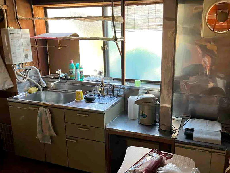 リフォーム前の古い木造住宅のキッチン。ステンレスシンク付きの流し台、壁面の瞬間湯沸かし器、換気扇、窓、調理器具が並ぶ、経年劣化が進んだ台所の様子。