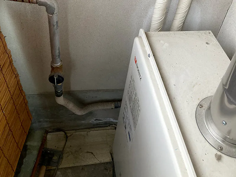 リフォーム前の室内に設置されたノーリツ製の古い給湯器ボイラー。コンクリート壁の隅に配置され、上部から排気筒や給水・給湯用の配管が接続されている交換前の状態。